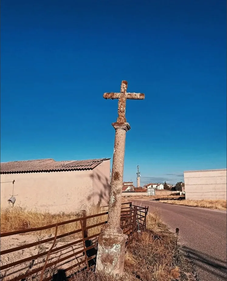 La Cruz del Canto, una cruz de piedra cubierta de líquenes sobre una columna lisa, junto a una cerca de ganado y una nave, con el camino asfaltado y el caserío de Cantalejo al fondo