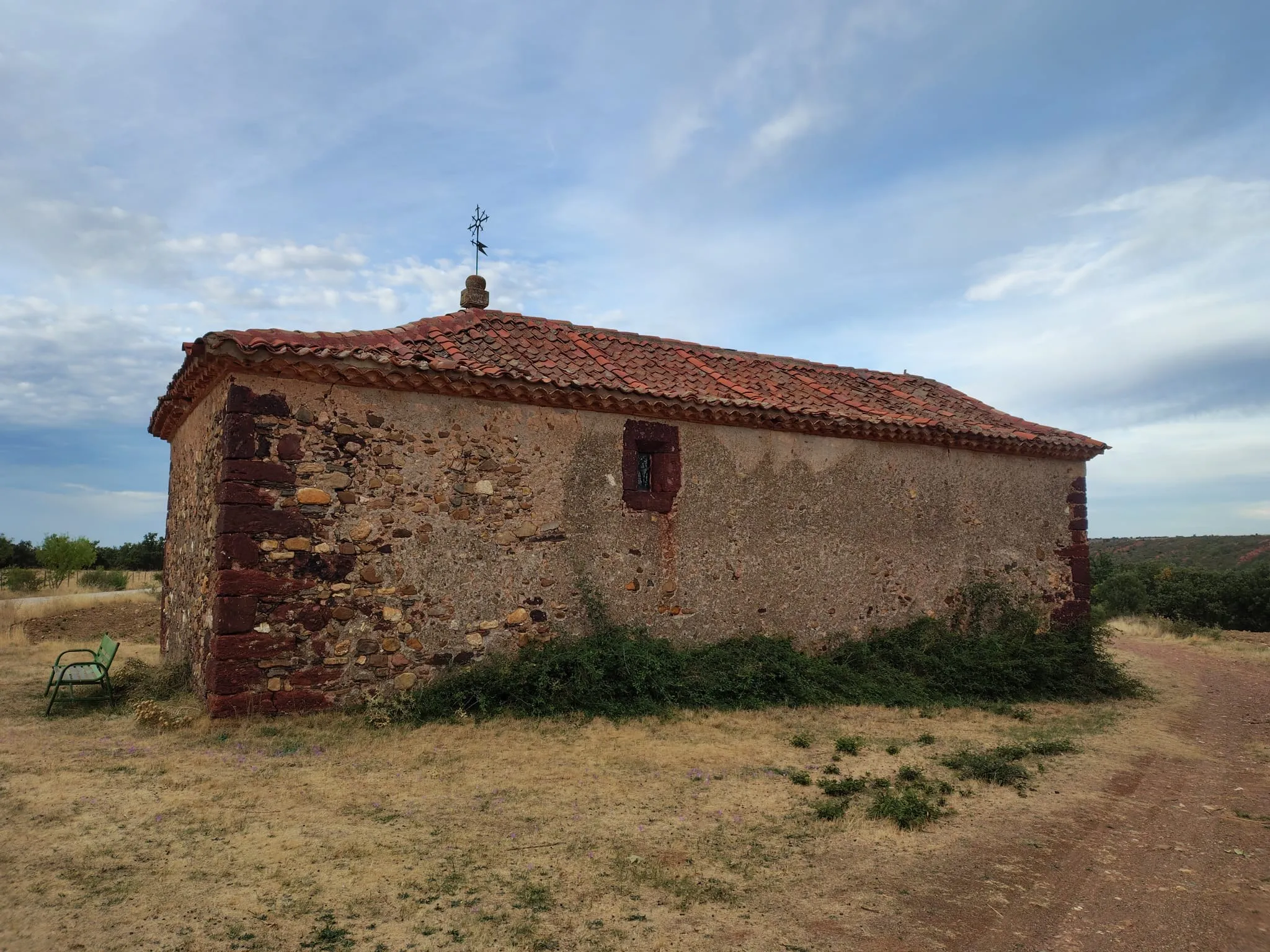 La ermita de San Roque de Villacorta desde el camino: muros de mampostería con las esquinas de rodeno, cubierta de teja curva a dos aguas y una cruz de forja sobre el tejado