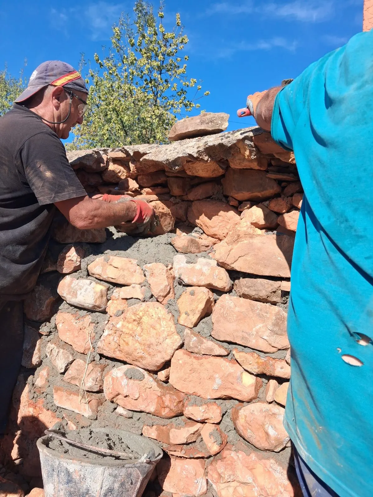 Un vecino con la paleta rellenando de mortero las juntas de la tapia, junto a otro que le acerca piedra