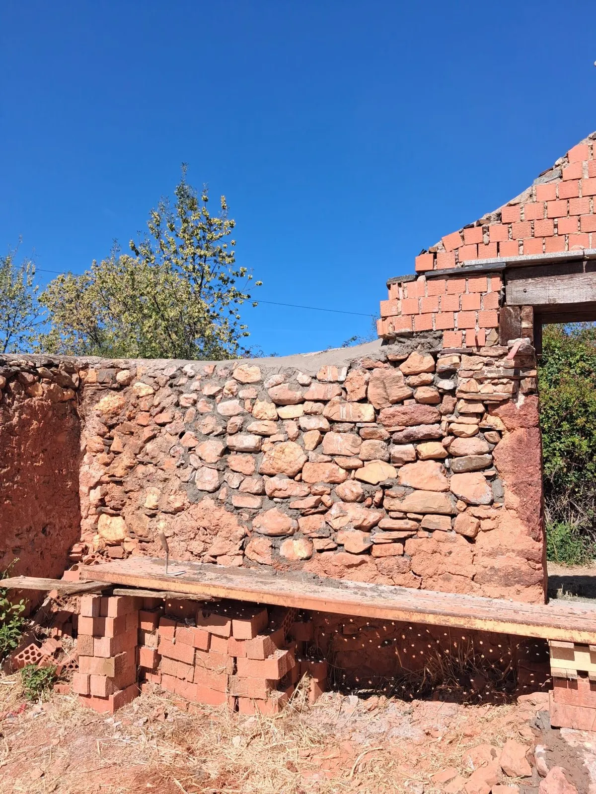 El tramo de la tapia del cementerio de Villacorta ya levantado, de piedra roja, con la portada de ladrillo al fondo y los tablones del andamio todavía al pie