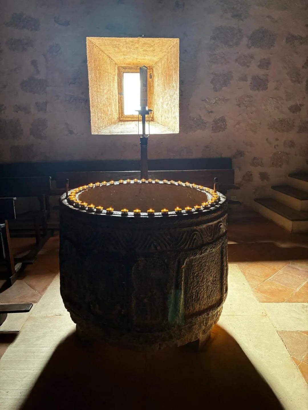La pila bautismal, cilíndrica y sin pie, iluminada a contraluz por una ventana de la nave, con la decoración de recuadros en relieve