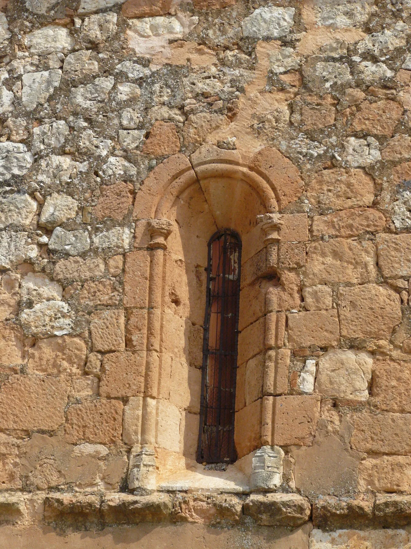 Una ventana estrecha de medio punto en un muro de sillería y mampostería rojiza, con una columnilla a cada lado rematada en capitel y una reja en el vano
