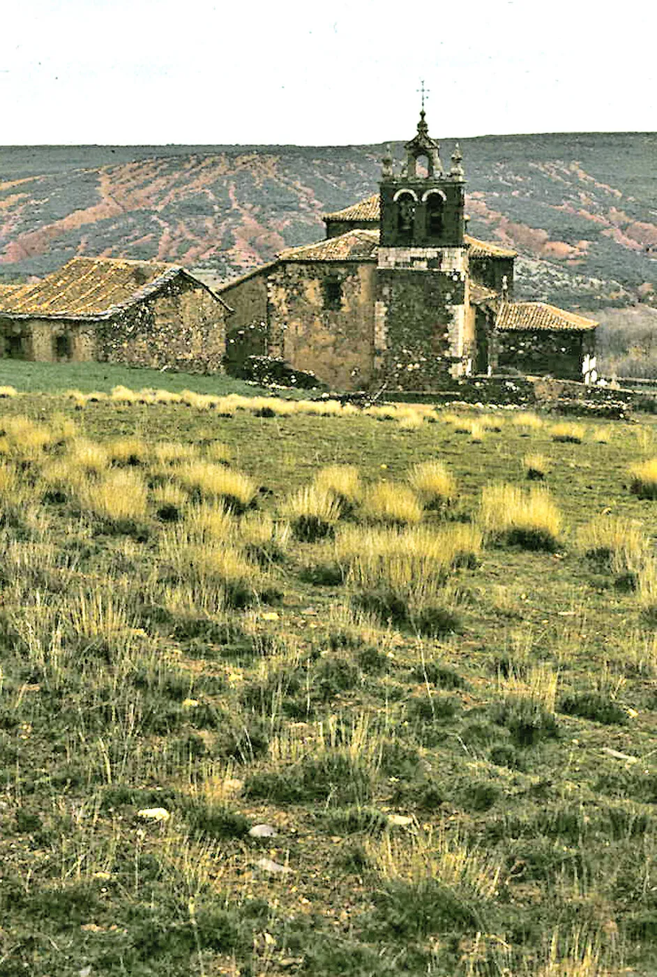 La iglesia de Madriguera y las casas de su alrededor, con un prado de hierba alta delante y los cerros rojos detrás, en 1974
