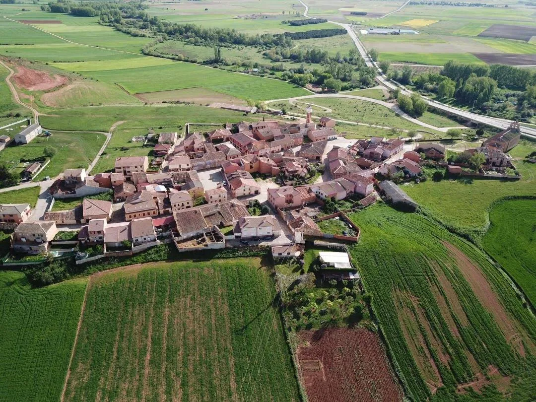 Santa María de Riaza desde el aire: el caserío de tejados rojos entre sembrados verdes, con la iglesia en el extremo de la terraza y la N-110 al fondo