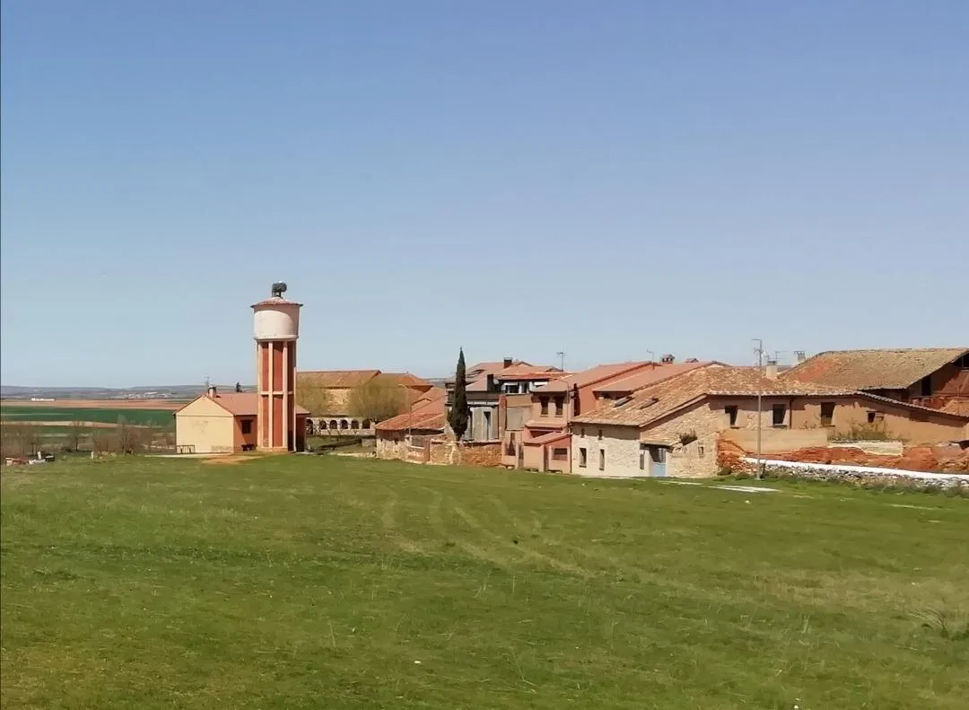 Casas de ladrillo y piedra rojiza junto a un prado, con el depósito de agua coronado por un nido de cigüeña y la llanura al fondo
