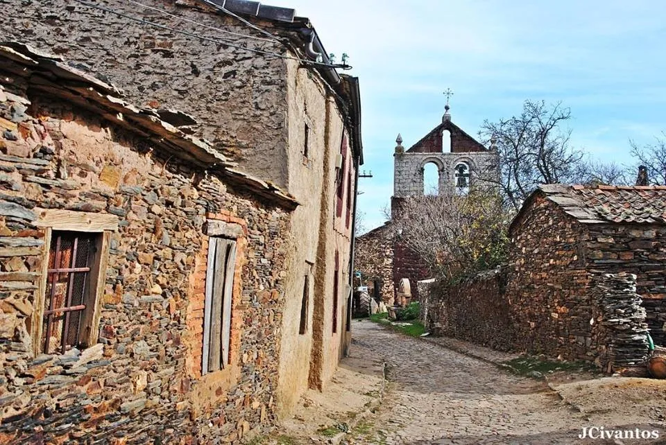 Calle empedrada de Serracín entre casas de pizarra oscura con esquinas y dinteles de rodeno rojo, con la espadaña de la iglesia al fondo