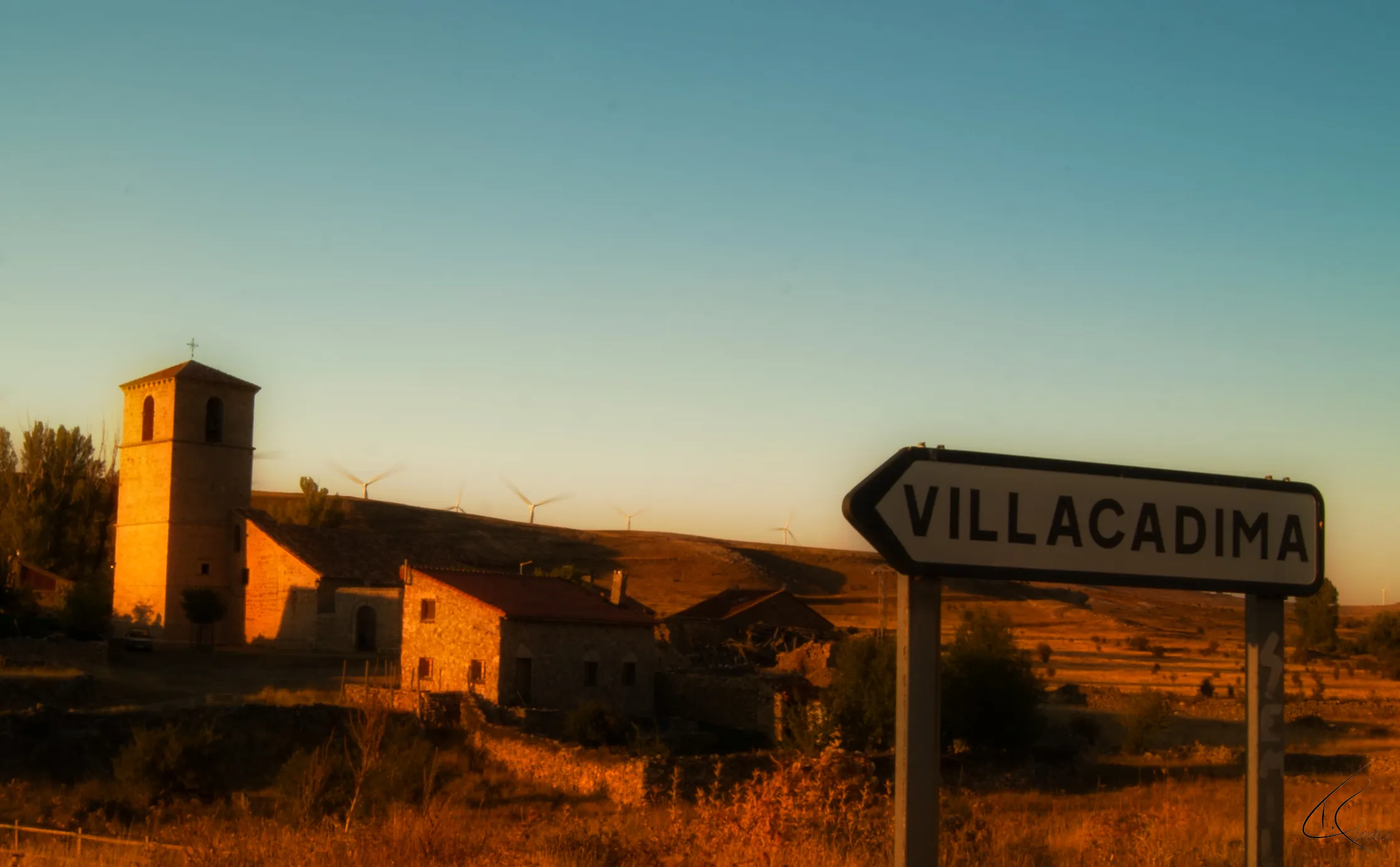 El cartel de Villacadima a la entrada del pueblo, con la torre de la iglesia y las casas al fondo a última hora de la tarde